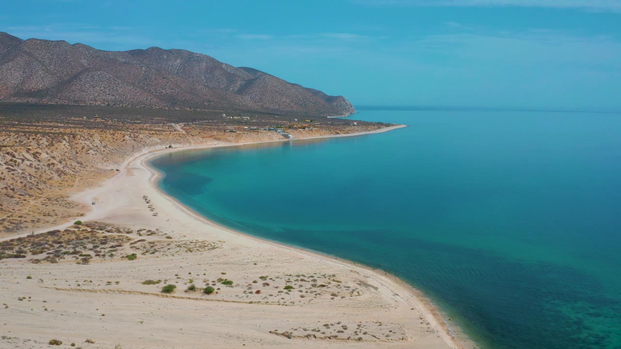 Aerial coastal frame with turquoise bay, light sand, and mountain backdrop.