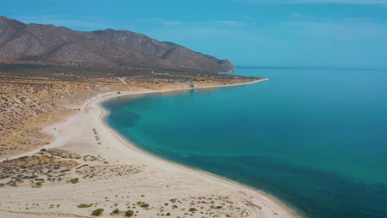 The second frame keeps the El Sargento La Ventana coastline wide, with the beach arc lower in frame, clear blue-green water filling the right side, mountains across the back, and no visible people.