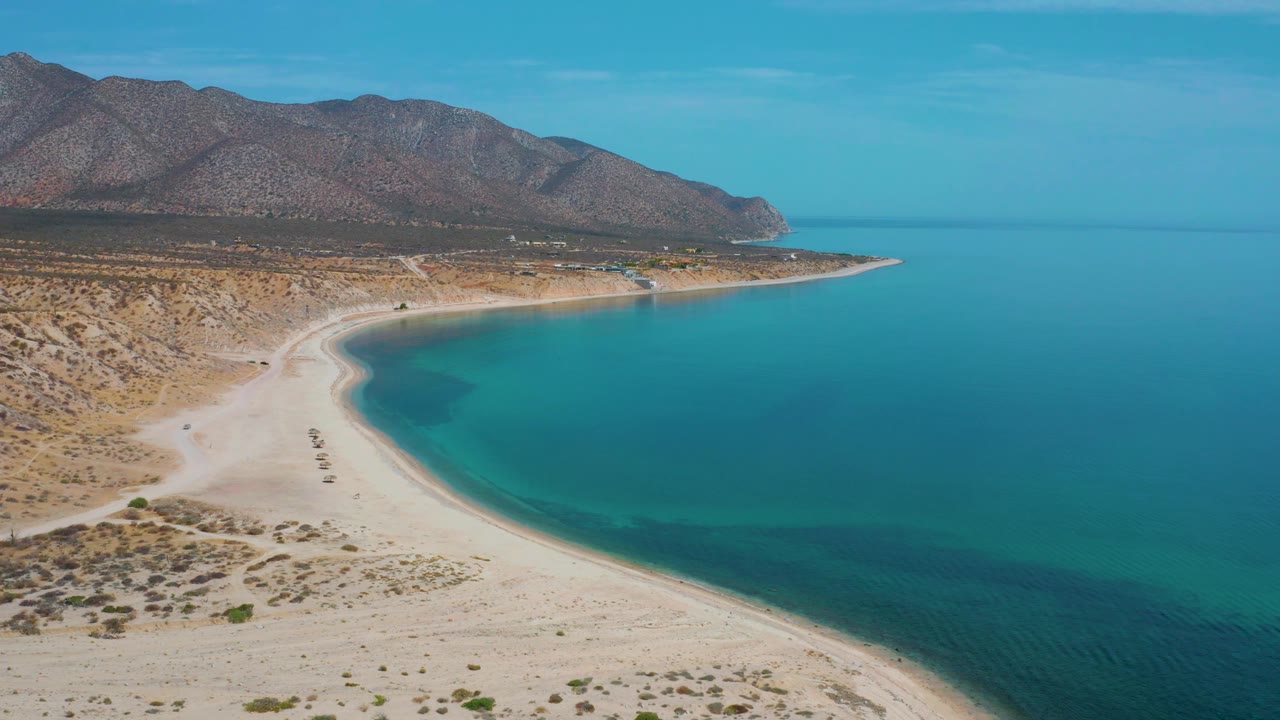 A later aerial frame shows the curved beach and shallow turquoise edge in El Sargento La Ventana, Mexico, with open blue sea to the right, rugged mountains behind, and no visible people.