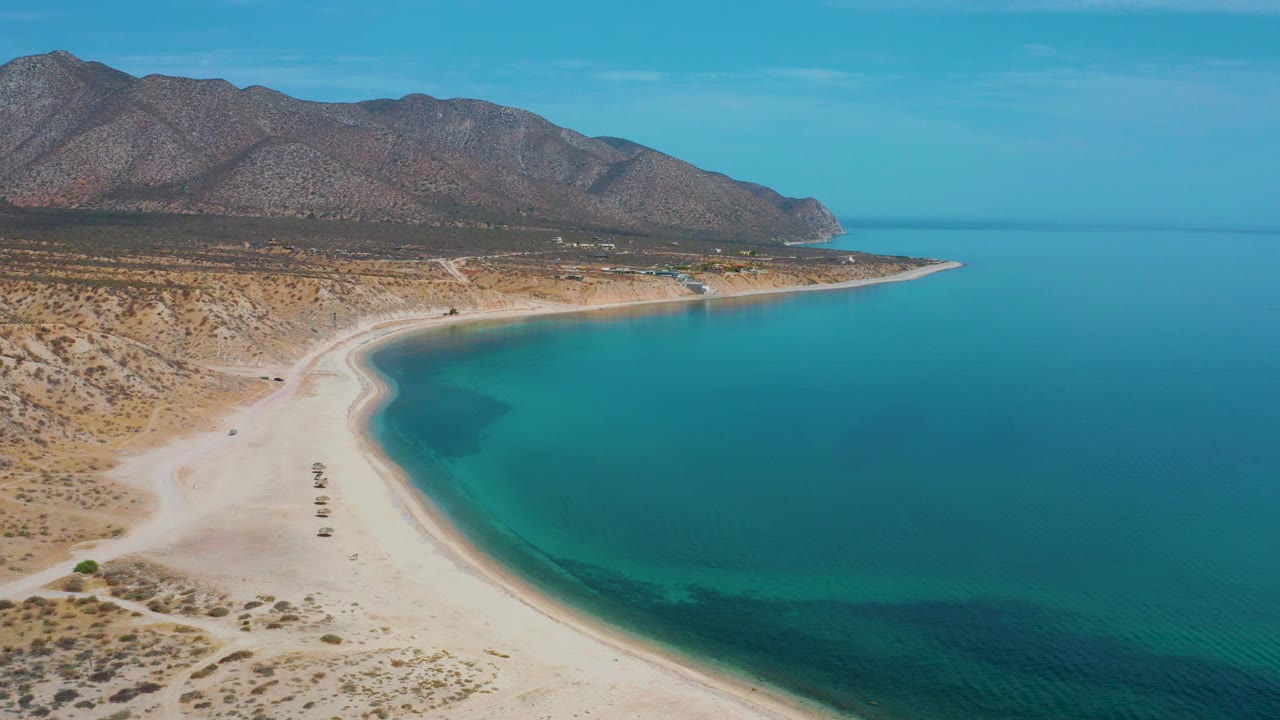 Opening aerial frame over El Sargento La Ventana, Mexico, with a pale curved beach on the left, blue-green sea on the right, desert scrubland, mountains, and no visible people.