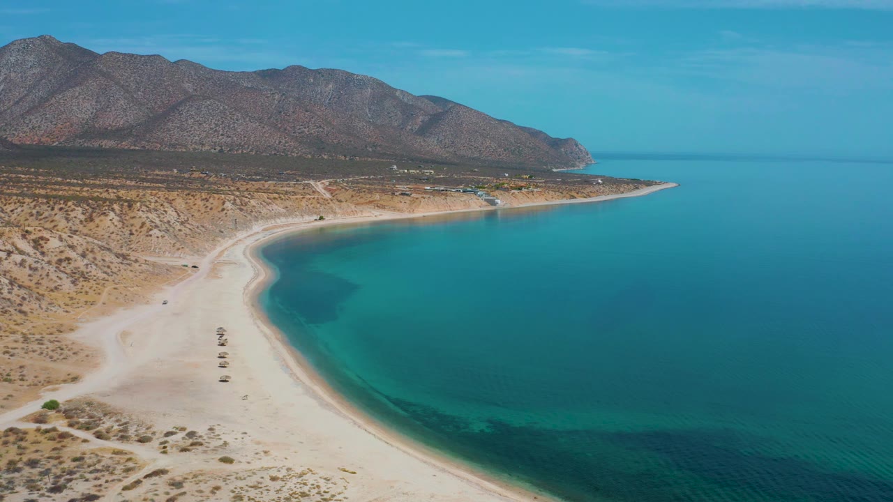 Aerial frame at 0:02 over El Sargento La Ventana, Mexico, with calm blue water occupying more of the right side, the beach bending through the center, arid hills behind it, and no visible people.