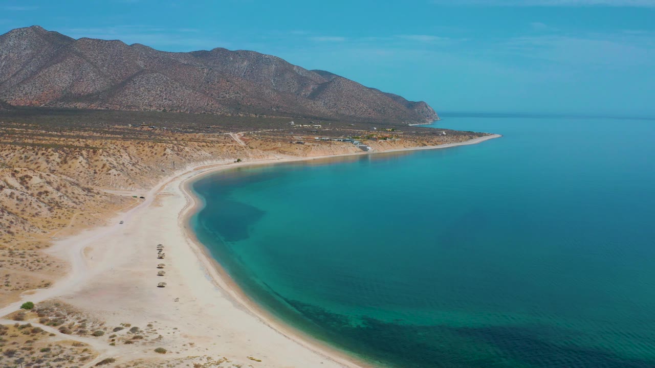 Aerial frame at 0:03 over El Sargento La Ventana, Mexico, showing more foreground sand, a narrow shoreline edge, blue water to the right, mountain slopes across the rear, and no visible people.