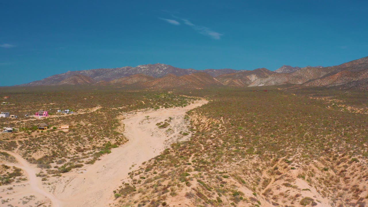 Aerial view in El Sargento La Ventana, Mexico, showing a pale sandy road through scrubland, small buildings at left, mountains on the horizon, and no visible people.
