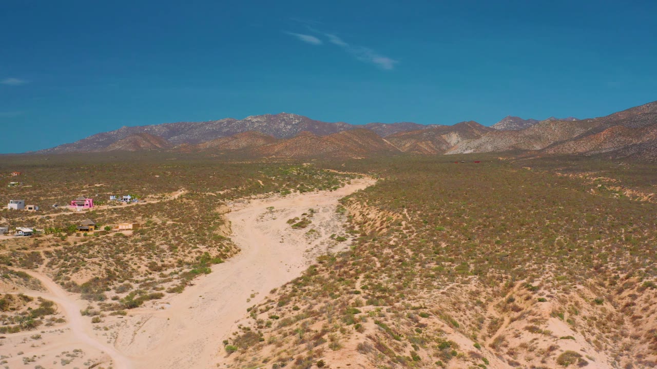 High aerial frame in El Sargento La Ventana, Mexico, looking over a pale road cutting through brushy terrain toward brown mountains, with small buildings at left and no visible people.