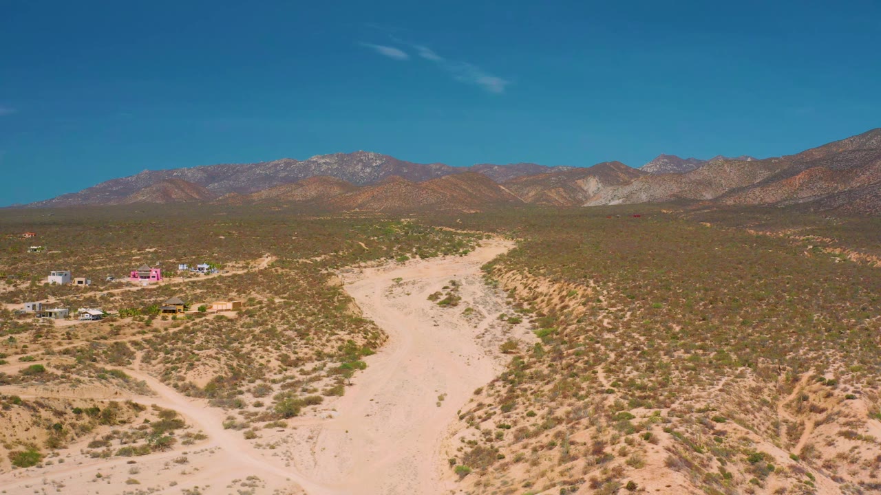 Wide desert aerial with pale road, scrub, small buildings, and mountains