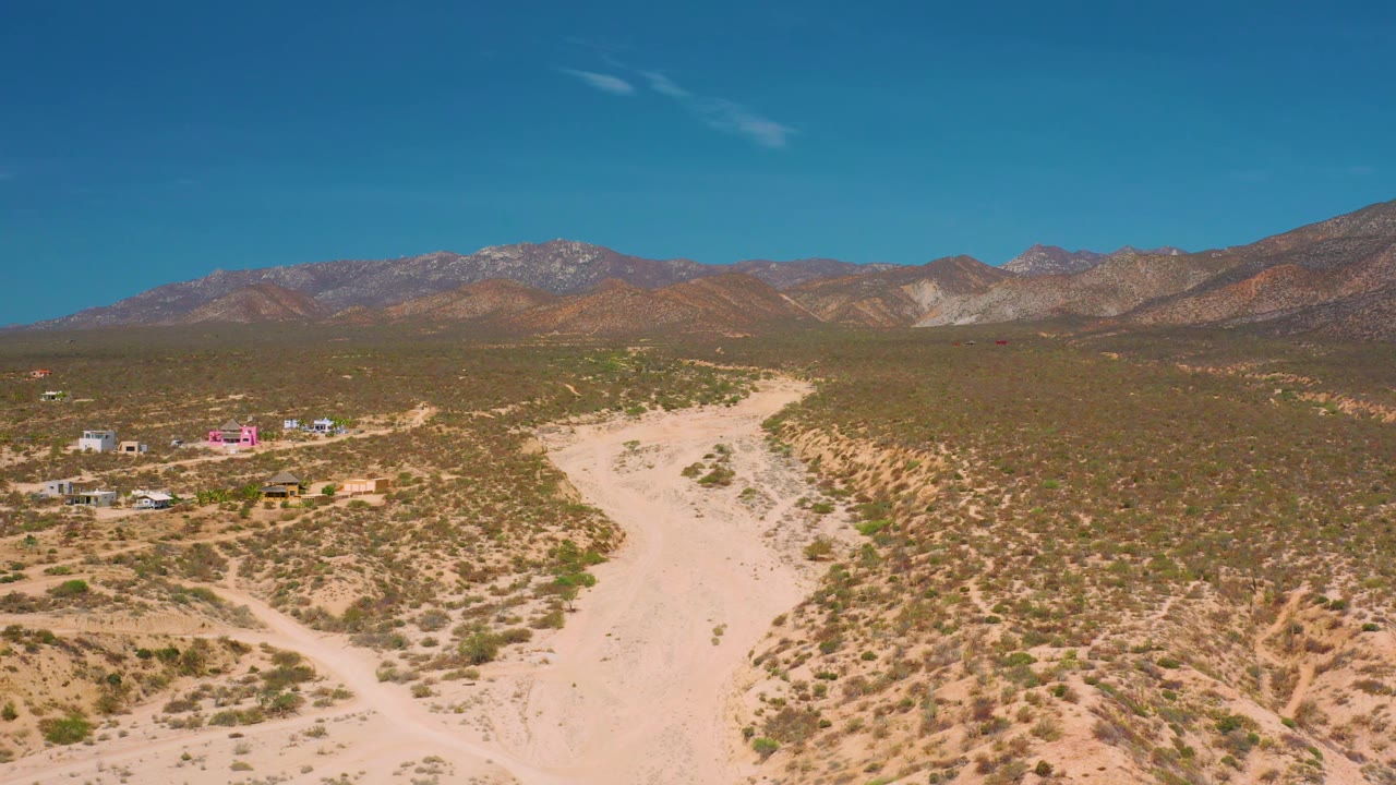 The frame looks across El Sargento La Ventana, Mexico scrubland toward brown mountains, with a winding sand track in the foreground and no visible people.