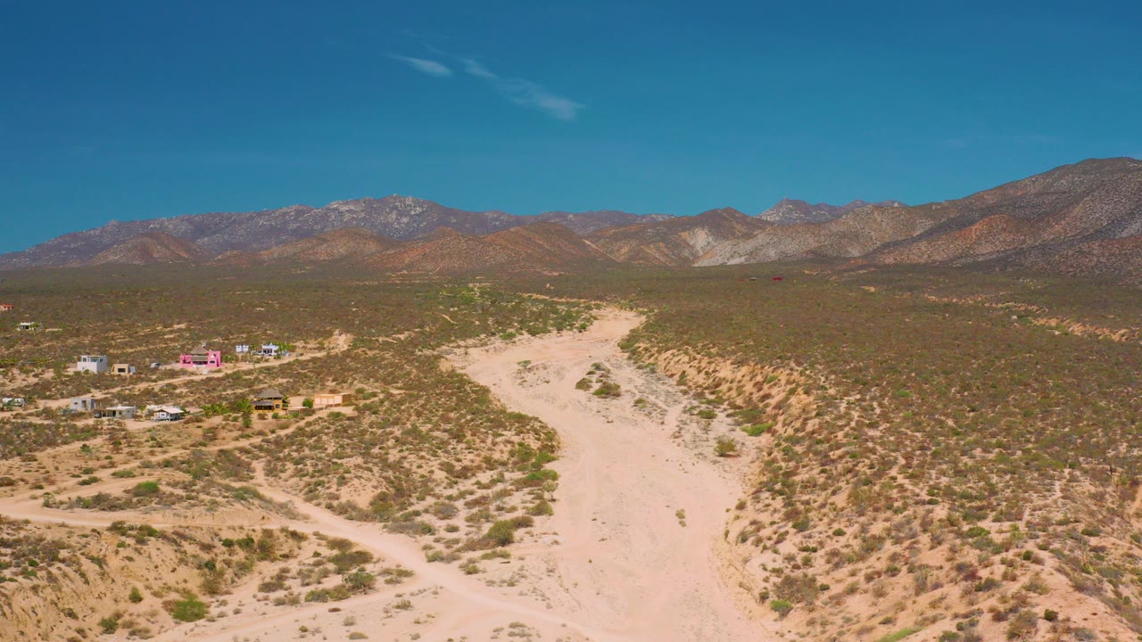 At 0:00, an elevated view in El Sargento La Ventana, Mexico shows a pale sandy wash, scrub-covered desert, small buildings to the left, distant mountains, blue sky, and no visible people.