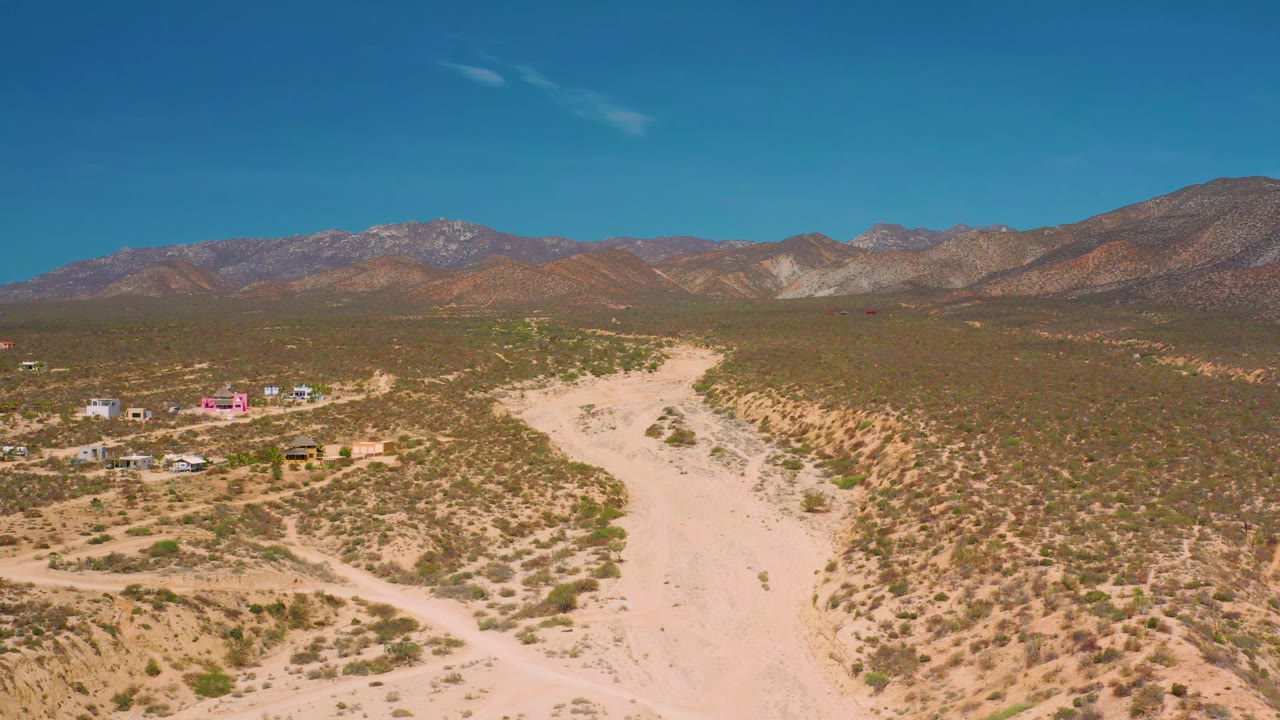 At 0:01, the aerial composition holds on a broad sandy channel through green-brown desert brush, with a small settlement at left, mountains across the horizon, and no visible people.