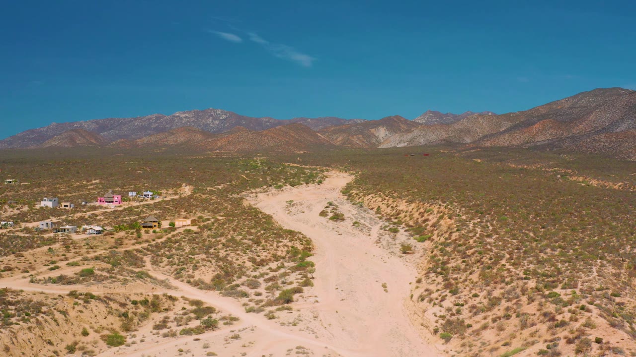 At 0:02, El Sargento La Ventana appears as a dry desert plain with a pale wash in the foreground, scattered buildings on the left edge, rugged mountains behind, and no visible people.
