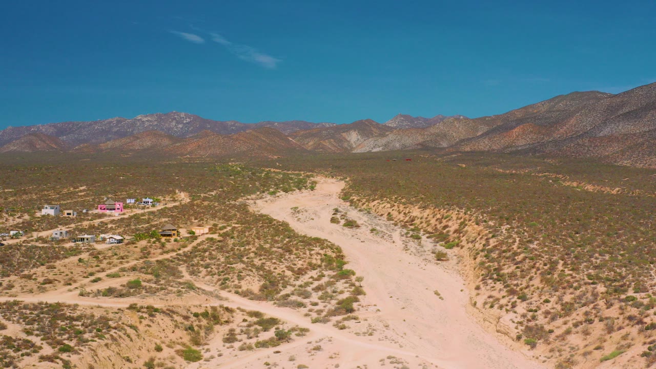 Opening aerial frame over El Sargento La Ventana, Mexico, showing a pale sandy wash, scattered desert shrubs, low buildings at left, distant mountains, blue sky, and no visible people.