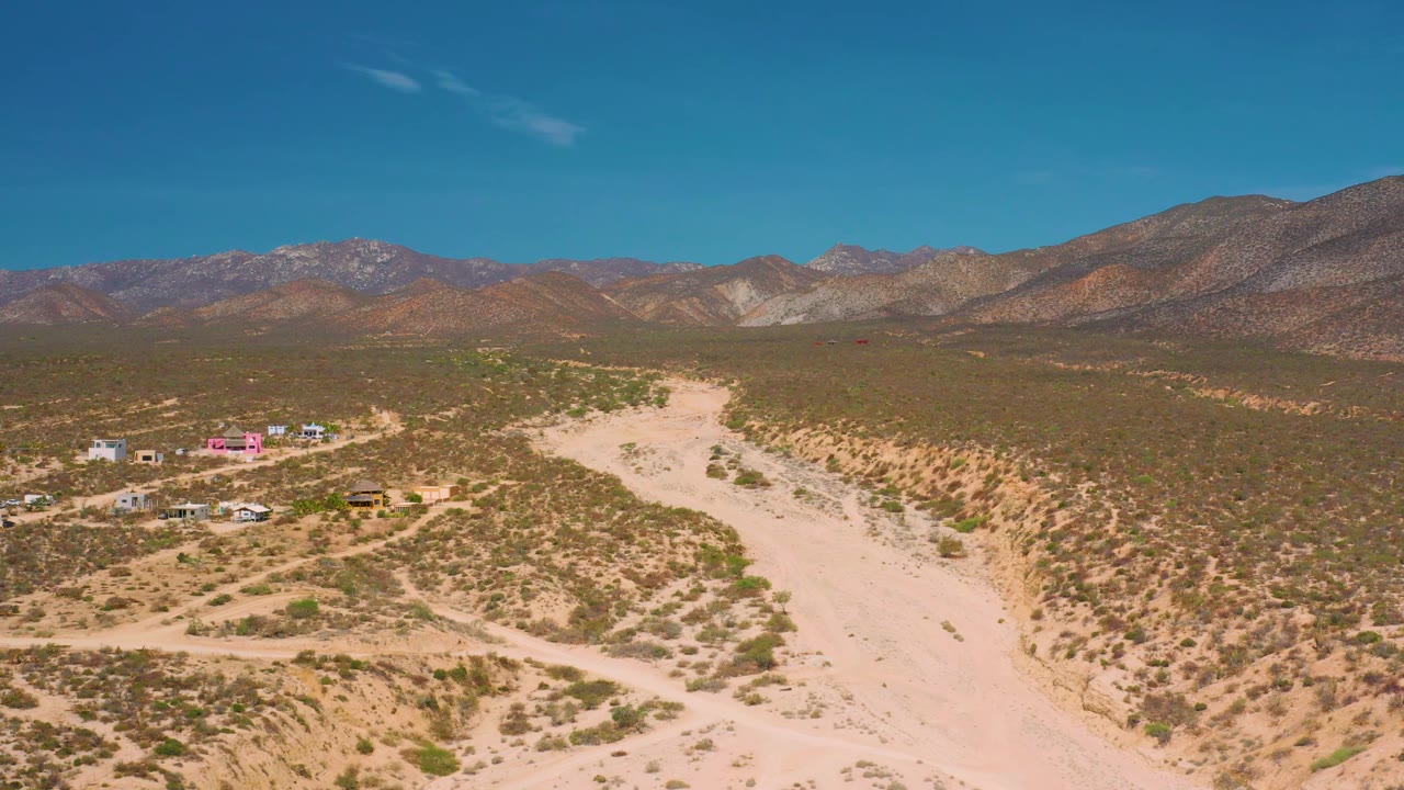 Wide elevated frame of El Sargento La Ventana, Mexico, showing a dry sandy channel through scrub, a few low buildings and vehicles to the left, layered mountains, and no visible people.