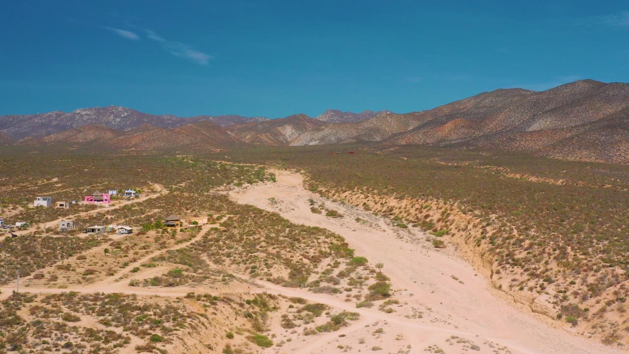Opening aerial frame over El Sargento La Ventana, Mexico, with a pale sandy wash curving through scrub desert, small buildings at left, mountain ridges beyond, and no visible people.