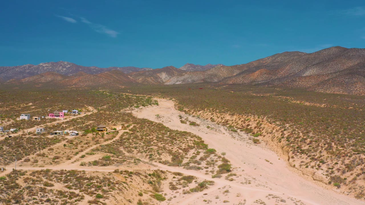 Aerial view over El Sargento La Ventana, Mexico, with the broad wash running from foreground toward midframe, sparse vegetation on both banks, small buildings at left, layered mountains, and no visible people.