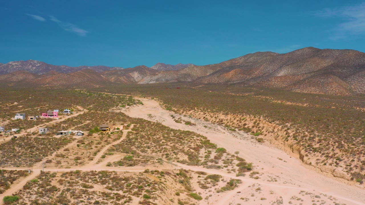 Wide aerial frame over El Sargento La Ventana, Mexico, with a pale dry river bed crossing the desert, small buildings at left, distant mountains, and no visible people.