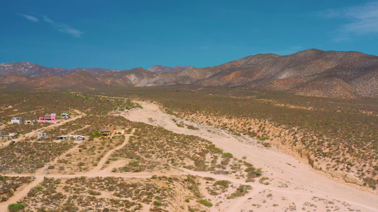 Drone-height frame in El Sargento La Ventana, Mexico, showing the pale dry wash widening across the foreground, scattered buildings to the left, mountains beyond, and no visible people.