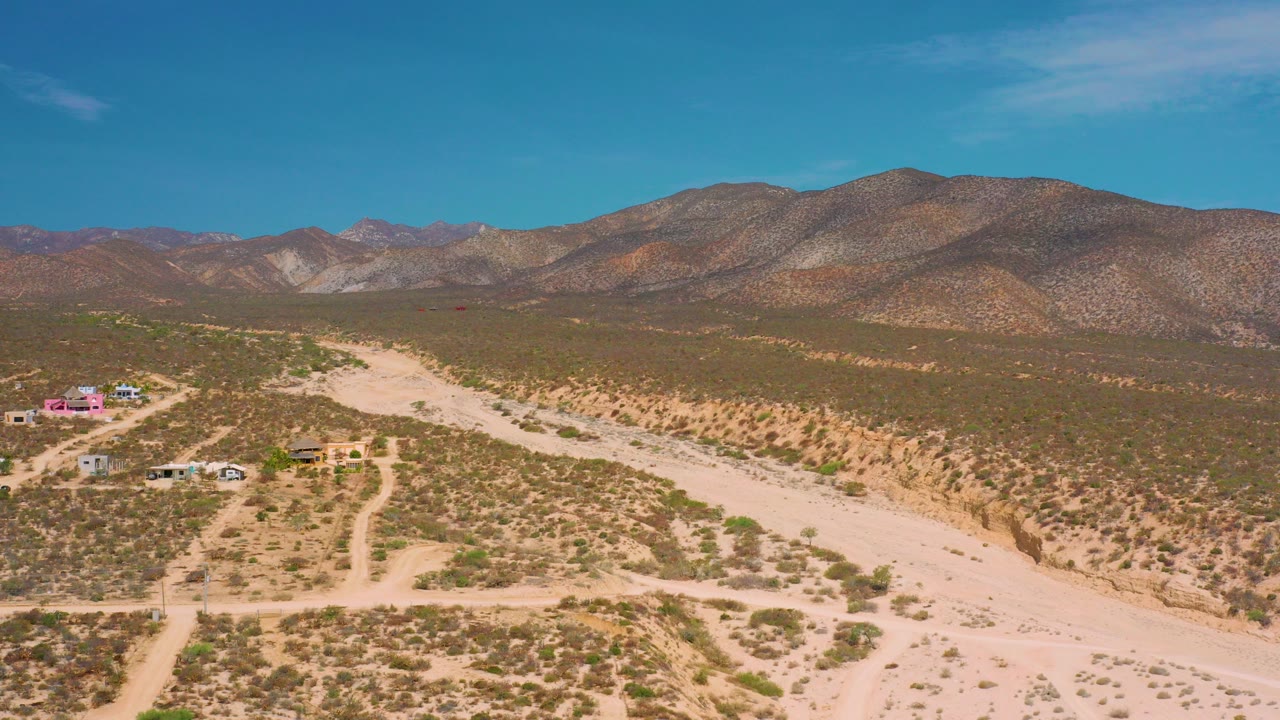 Opening aerial frame over El Sargento La Ventana, Mexico, with a pale sandy wash cutting through scrub desert, a few small buildings at left, mountains on the horizon, and no visible people.