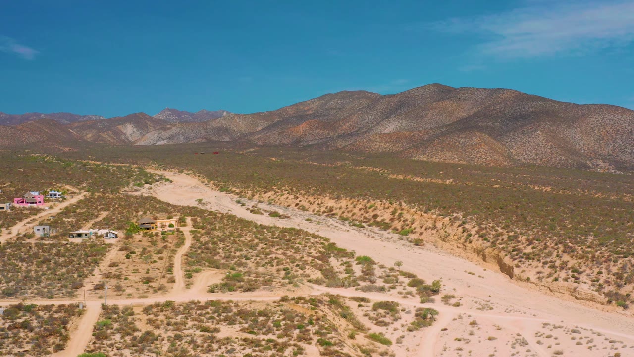 Aerial frame over El Sargento La Ventana, Mexico, with the dry wash running from lower right toward the middle distance, scattered shrubs, small buildings to the left, and no visible people.