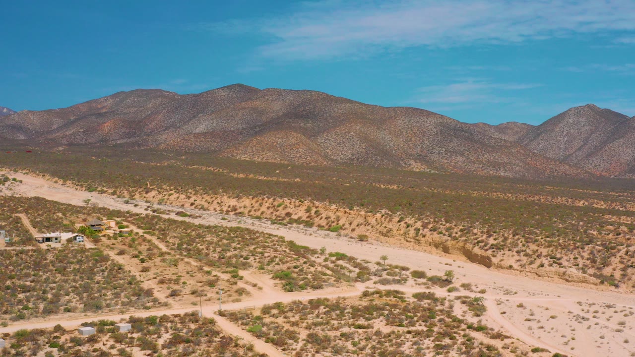 Opening frame over El Sargento La Ventana desert scrub, with a pale dirt road, small structures at left, distant mountains, blue sky, and no visible people.