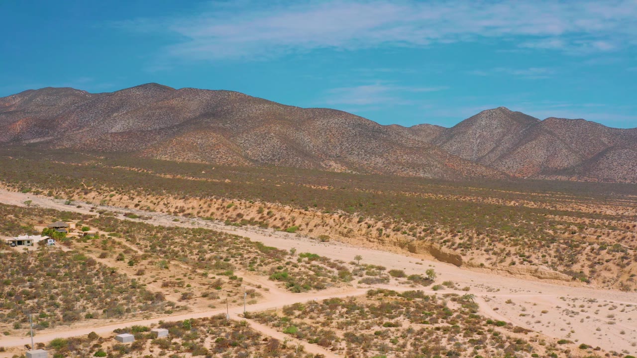 Wide frame in El Sargento La Ventana, Mexico, showing a desert valley of tan soil and green-gray scrub, rounded mountains in the background, and no visible people.
