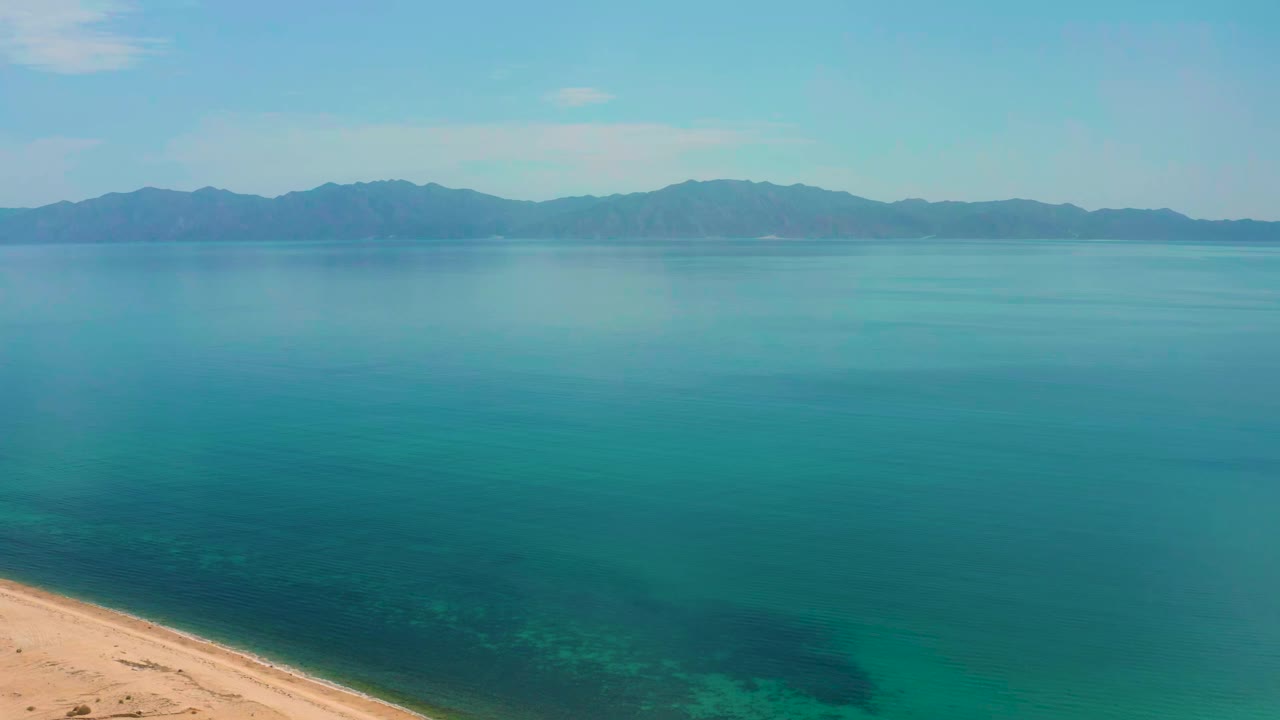 Opening frame over El Sargento La Ventana, Mexico with turquoise water across the view, sandy shore entering at lower left, distant mountains on the horizon, and no visible people.