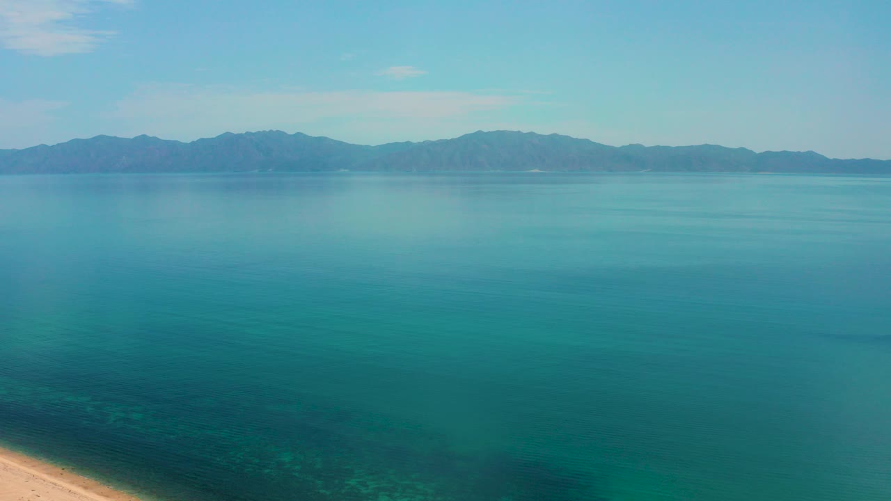 Wide aerial view over turquoise water in El Sargento La Ventana, Mexico, with distant mountains on the horizon, a small sandy shoreline edge at lower left, and no visible people.
