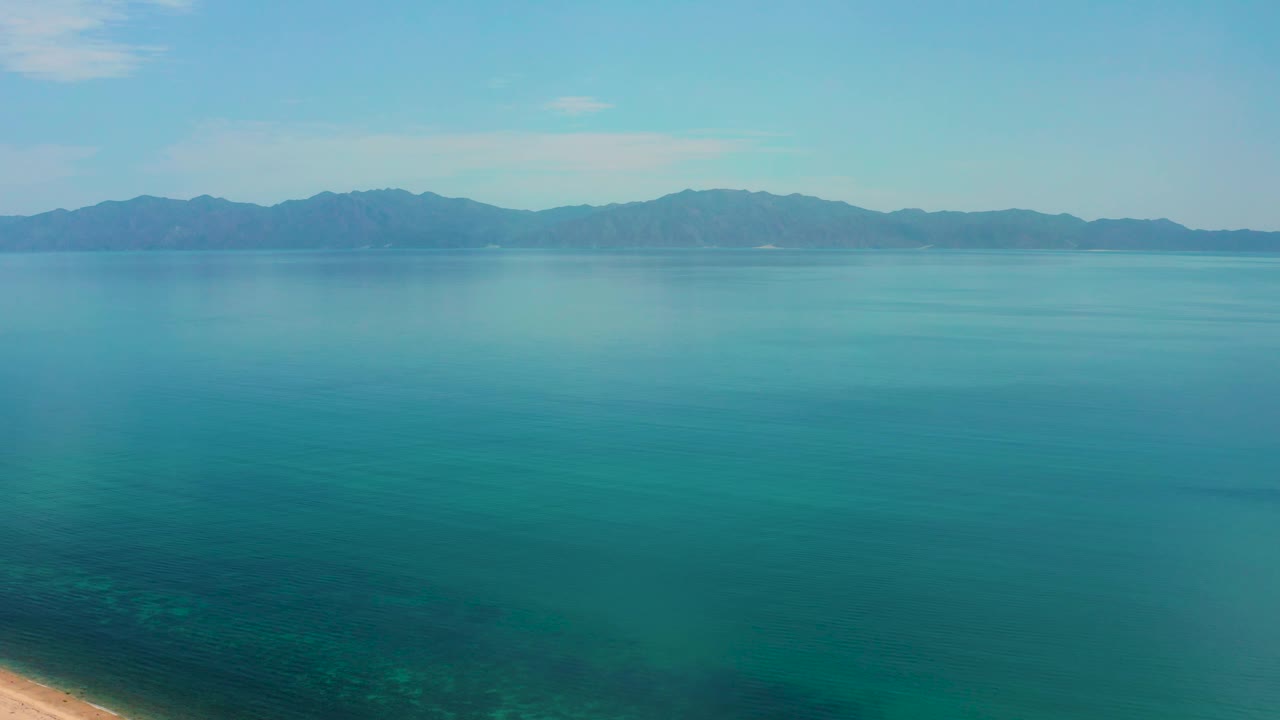 High view in El Sargento La Ventana, Mexico, with blue-green sea dominating the frame, a thin beach sliver near the lower left edge, distant mountains, and no visible people.