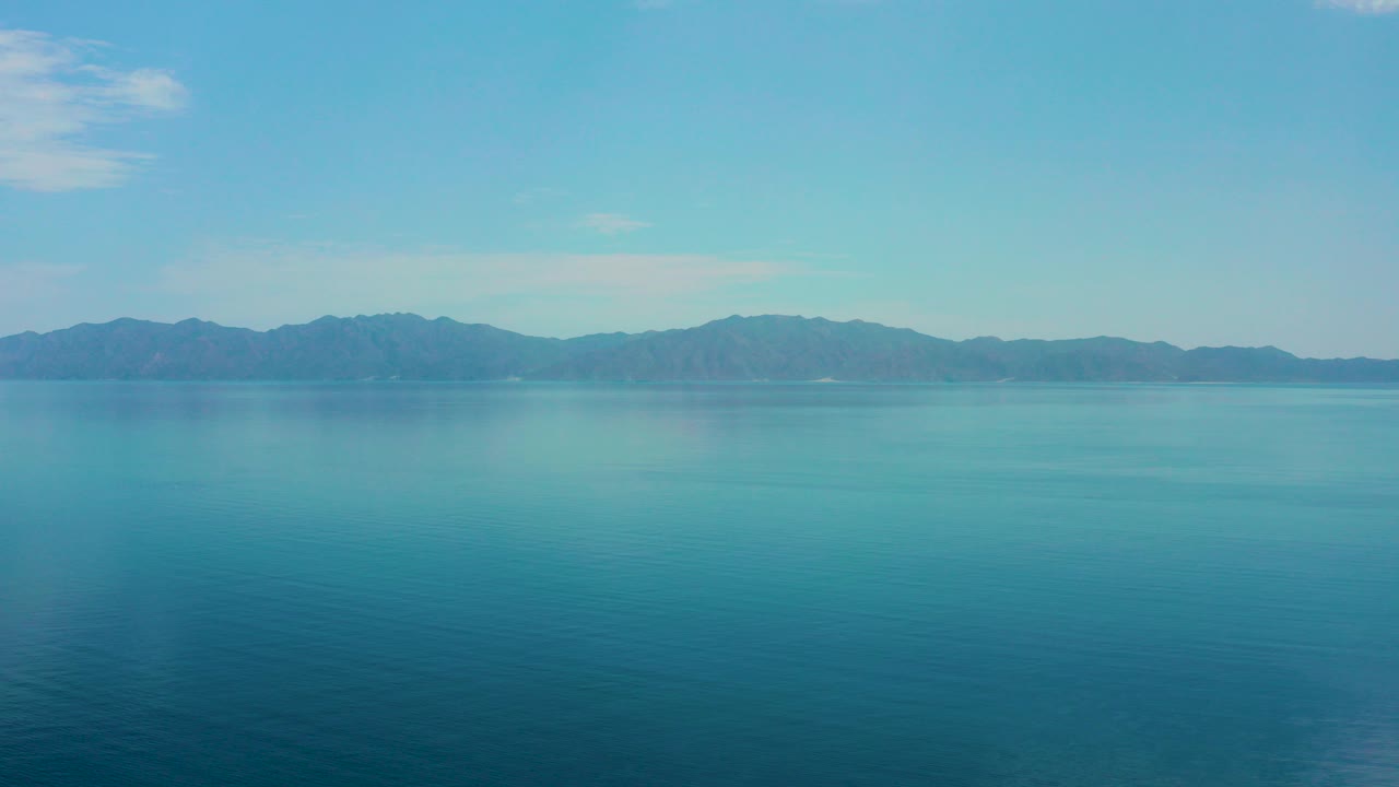 Wide view of blue sea and a distant mountain ridge in El Sargento La Ventana, Mexico, with pale sky above and no visible people.