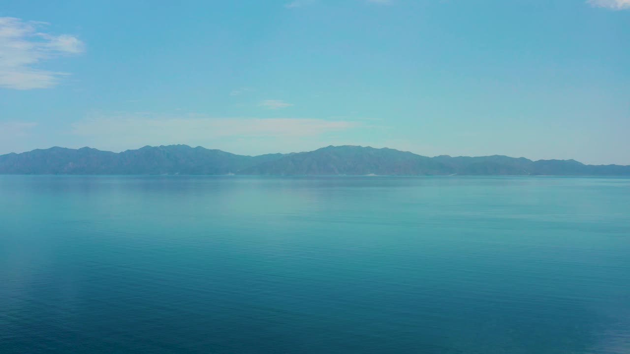 Open blue ocean sits below a long mountain ridge in El Sargento La Ventana, Mexico, with small white clouds high in the frame and no visible people.