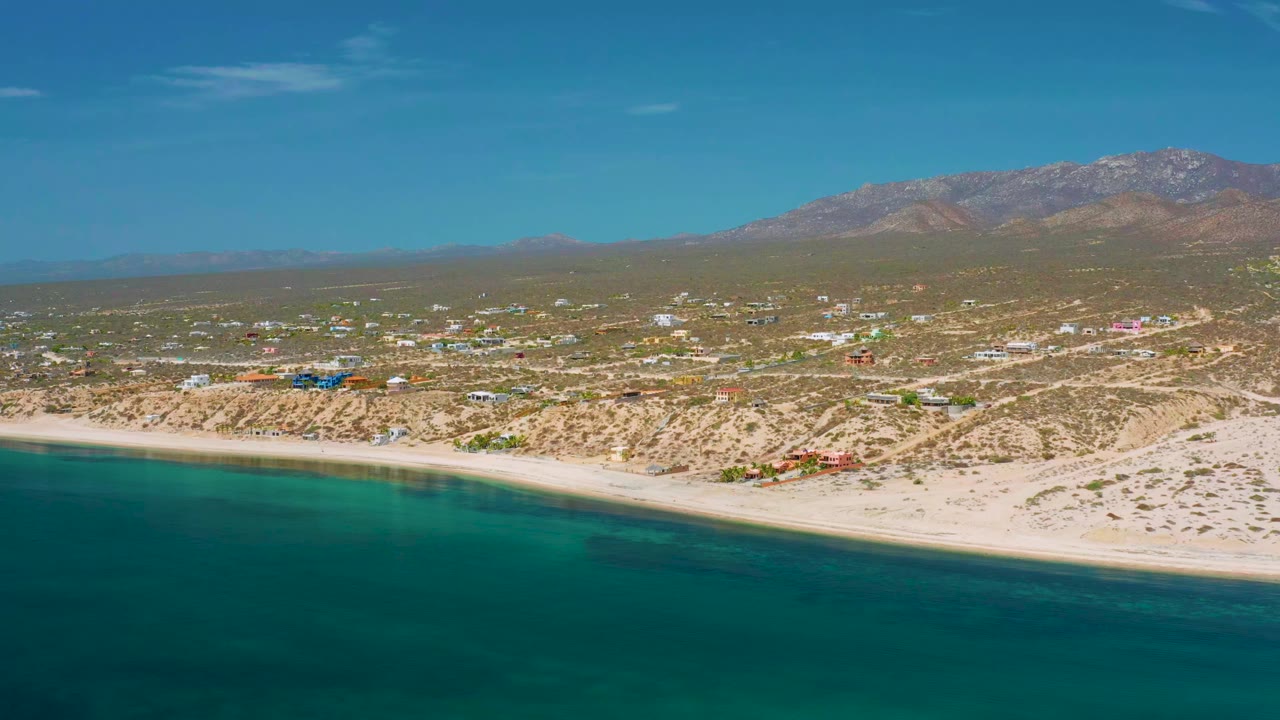Wide elevated view of El Sargento La Ventana, Mexico, showing calm teal water, a long sandy shoreline, scattered low buildings, dry terrain, distant mountains, and no visible people.
