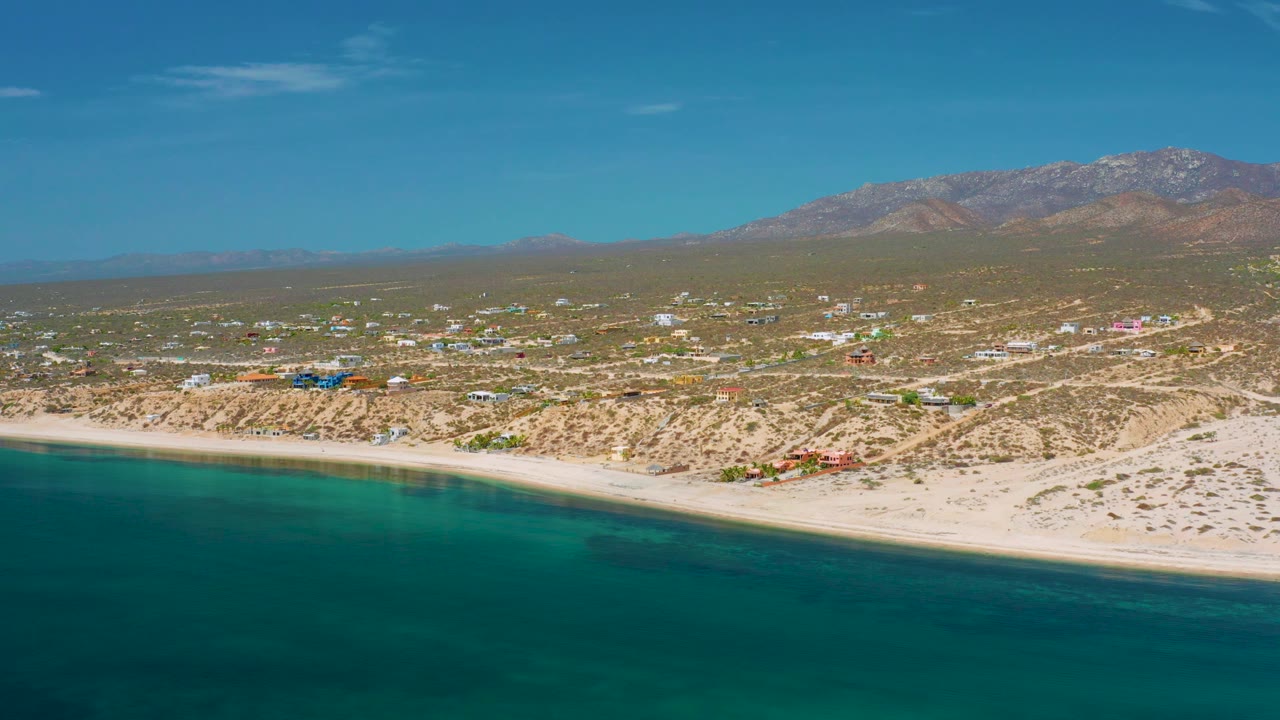 Elevated aerial frame in El Sargento La Ventana, Mexico, with blue-green sea across the lower frame, a bright sand strip, small structures inland, a mountain ridge at the horizon, and no visible people.