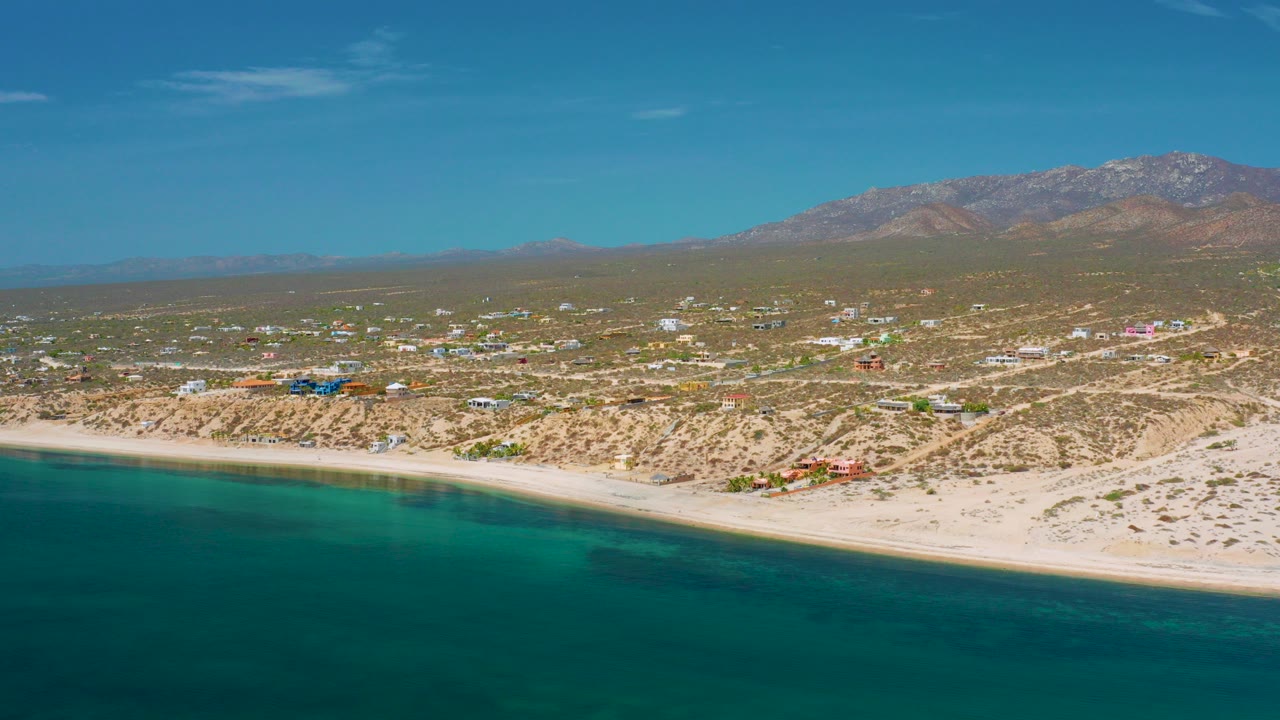 Wide aerial view over El Sargento La Ventana, Mexico, with teal water in the foreground, pale beach, scattered town buildings, dry terrain, distant mountains, and no visible people.