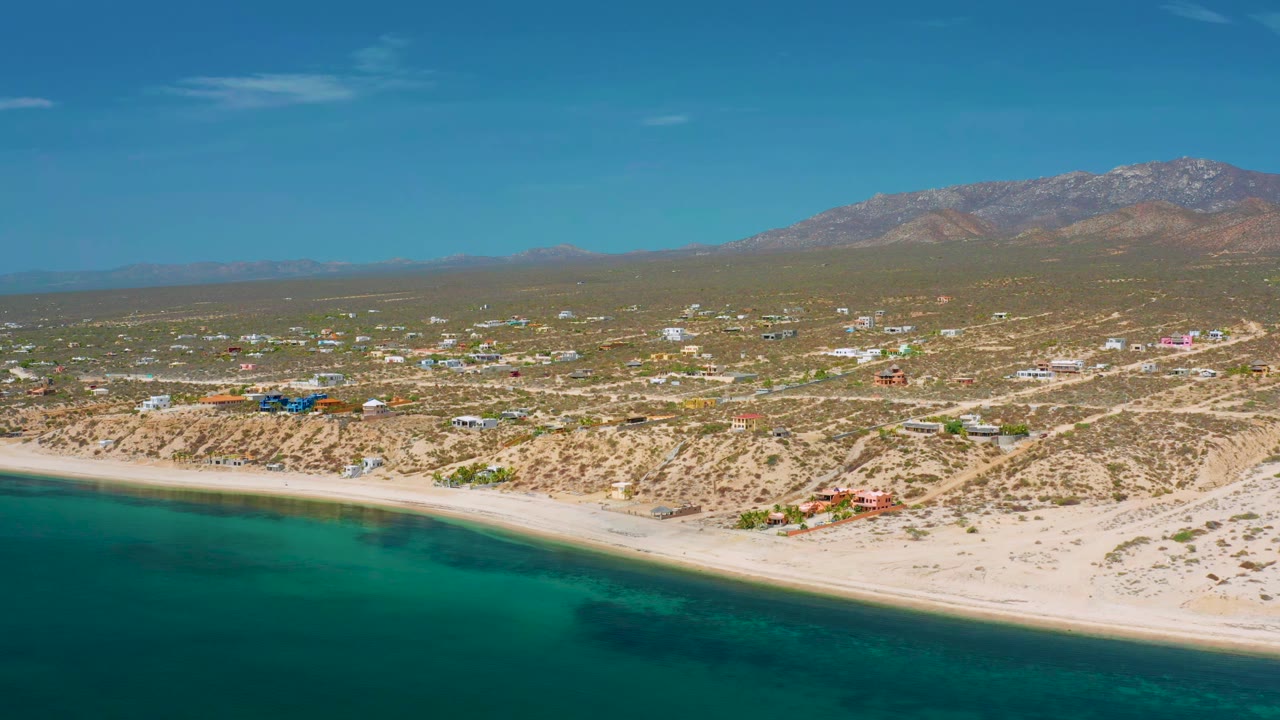 Aerial frame over El Sargento La Ventana showing a longer run of shoreline, darker water patches near the beach, desert roads, low buildings, mountains, and no visible people.
