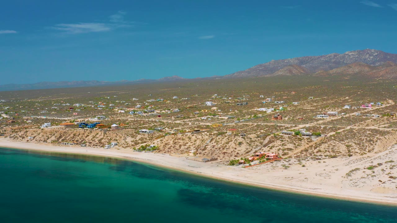 A wide aerial frame of El Sargento La Ventana with the shoreline crossing the lower frame, scattered desert structures across the midground, a broad mountain backdrop, and no visible people.