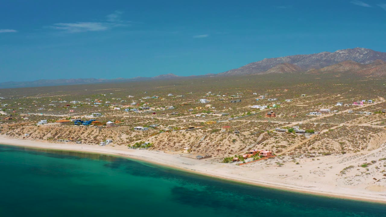 Aerial shoreline with calm sea, sandy beach, desert roads, buildings, and mountains.