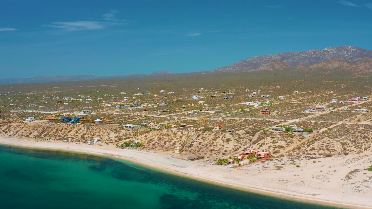 Aerial frame over El Sargento La Ventana, Mexico with turquoise water at the lower left, a pale beach, scattered desert buildings, dirt roads, distant mountains, and no visible people.