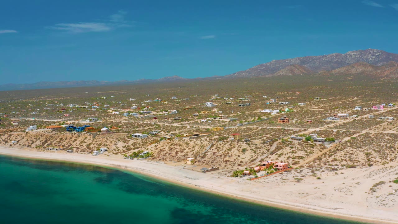Wide aerial coastline with beach, desert lots, scattered buildings, and mountains