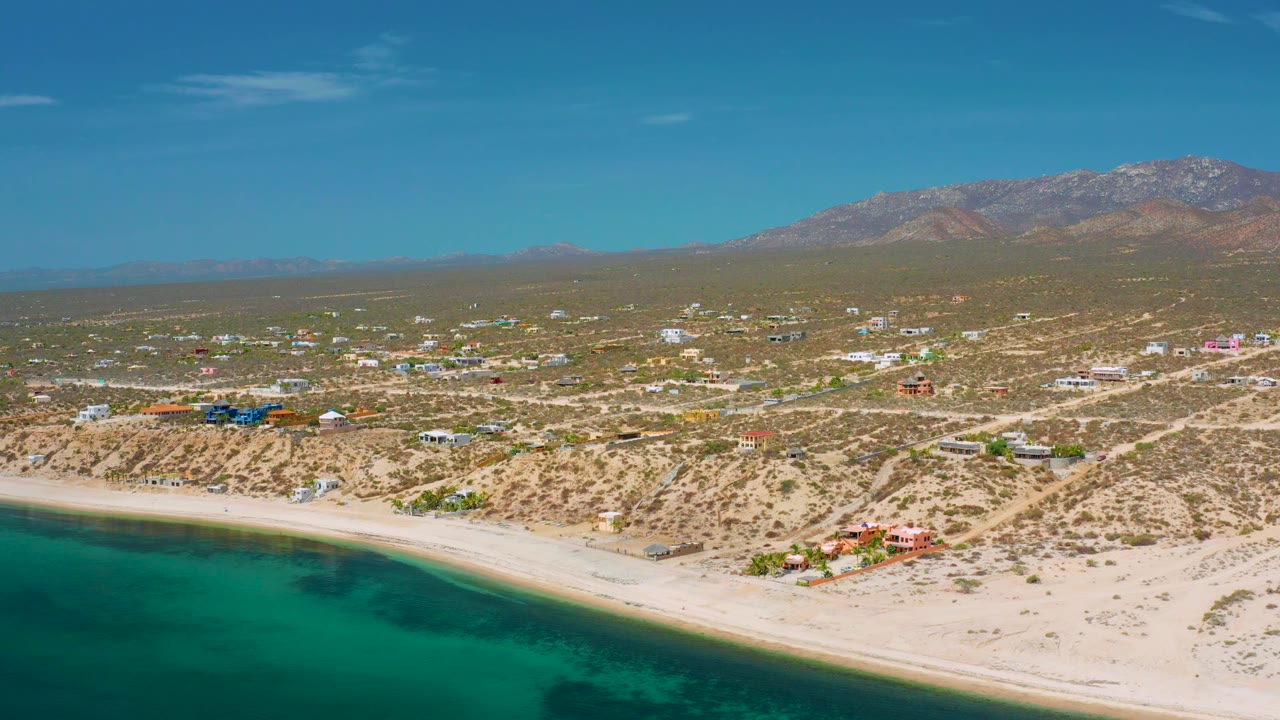 Aerial view across El Sargento La Ventana, Mexico with turquoise sea in the foreground, a bright sand beach, scattered desert homes and roads, hazy mountain background, and no visible people.