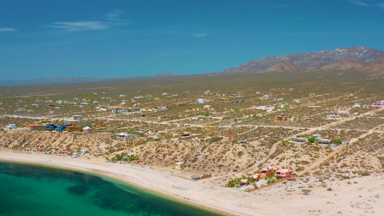 Wide aerial frame over El Sargento La Ventana, Mexico, with turquoise water at lower left, pale sand, scattered homes, dry roads, distant mountains, and no visible people.