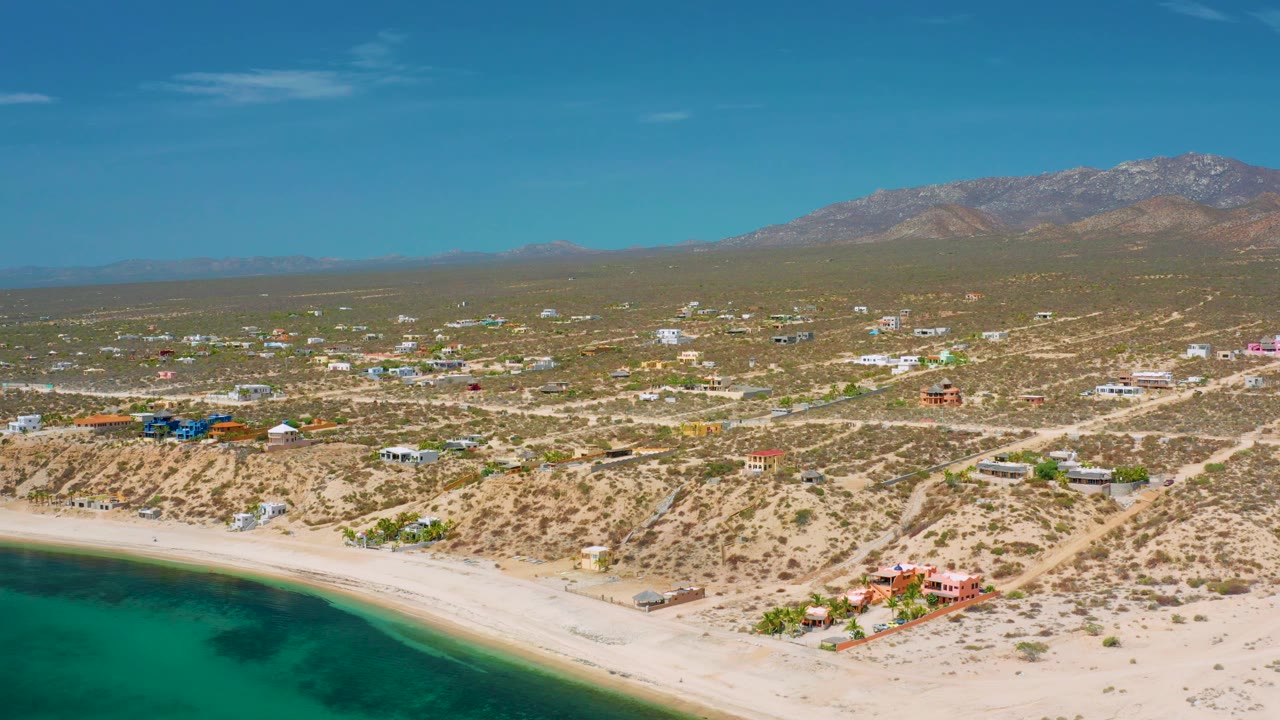 El Sargento La Ventana appears from above with pale beach at the bottom, scattered residential buildings across dry lots, a broad mountain backdrop, and no visible people.