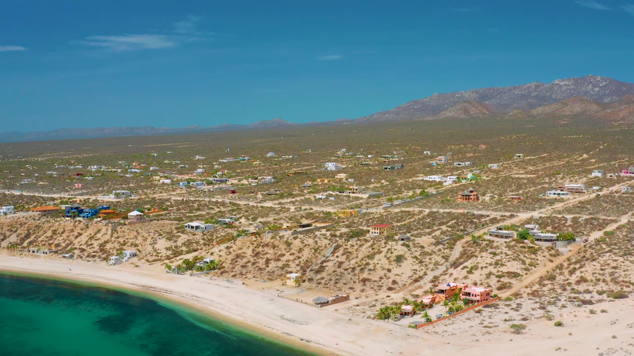 High coastal view with turquoise water, sand, desert homes, and mountain horizon.
