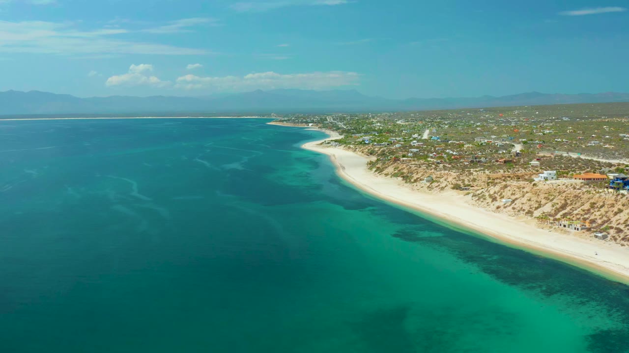 Aerial frame over El Sargento La Ventana, Mexico, showing deeper teal water at left, lighter shallows near the beach, a diagonal sand strip, low vegetation, scattered structures, and no visible people.