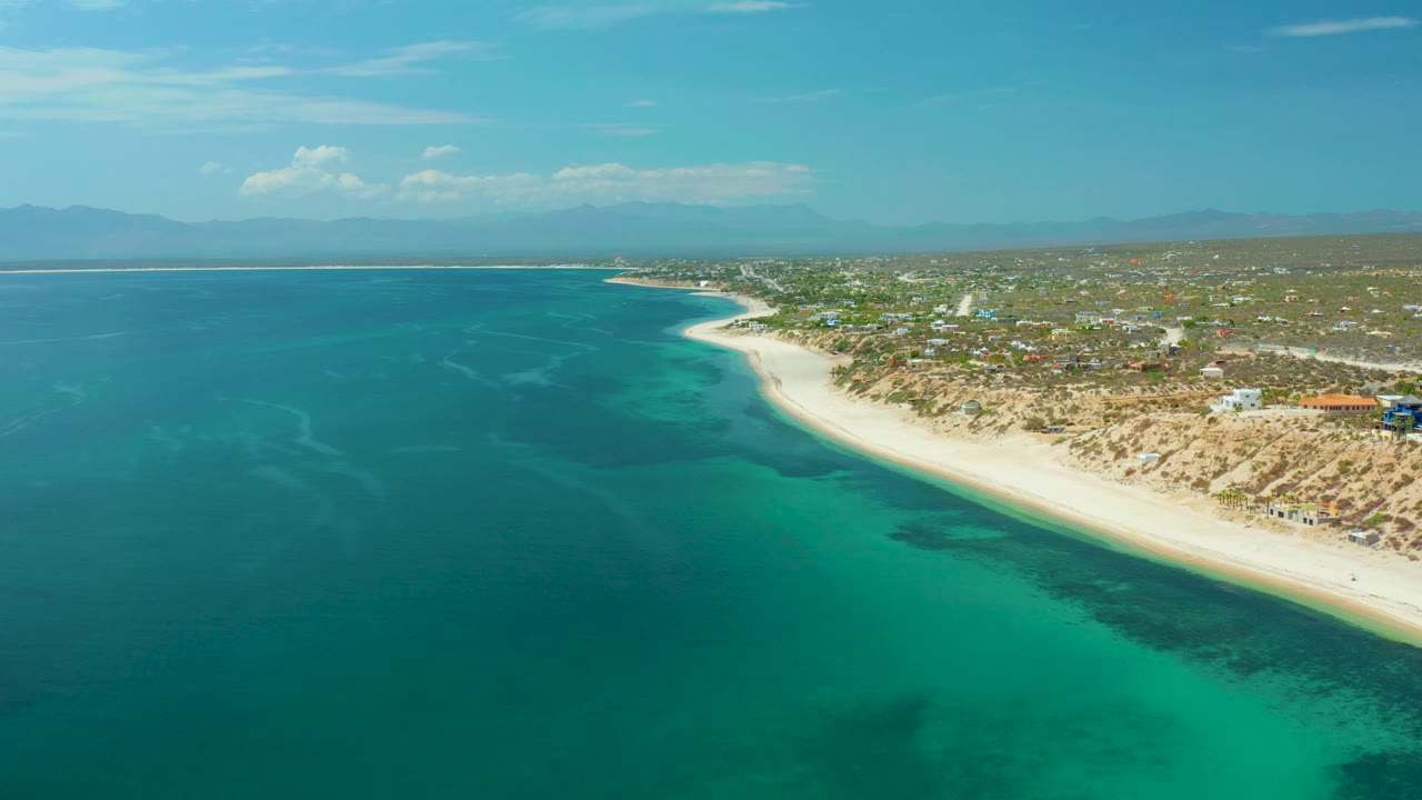 Aerial coastline with teal water, pale beach, inland vegetation, and distant mountains.