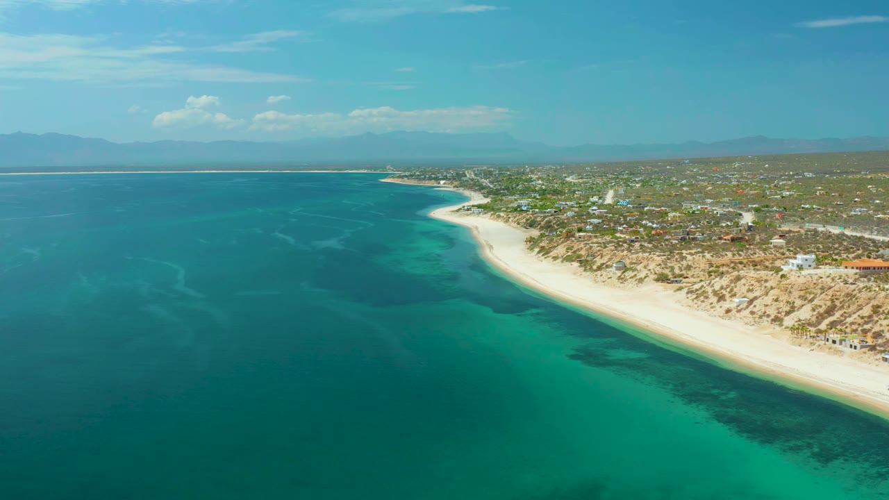 A wide aerial frame in El Sargento La Ventana shows dark blue and green water patterns along the left, a bright sandy beach sweeping rightward, scattered coastal structures inland, and no visible people.