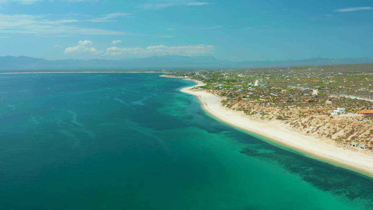 The frame holds a high coastal view of El Sargento La Ventana, with clear teal water in the foreground, darker wave and seabed shapes near shore, pale sand, inland brush, and no visible people.