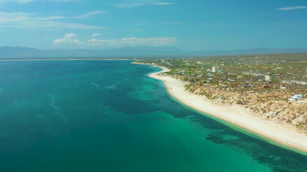 A high aerial view over El Sargento La Ventana, Mexico, shows turquoise water meeting a pale sandy beach with scattered inland buildings and no visible people.