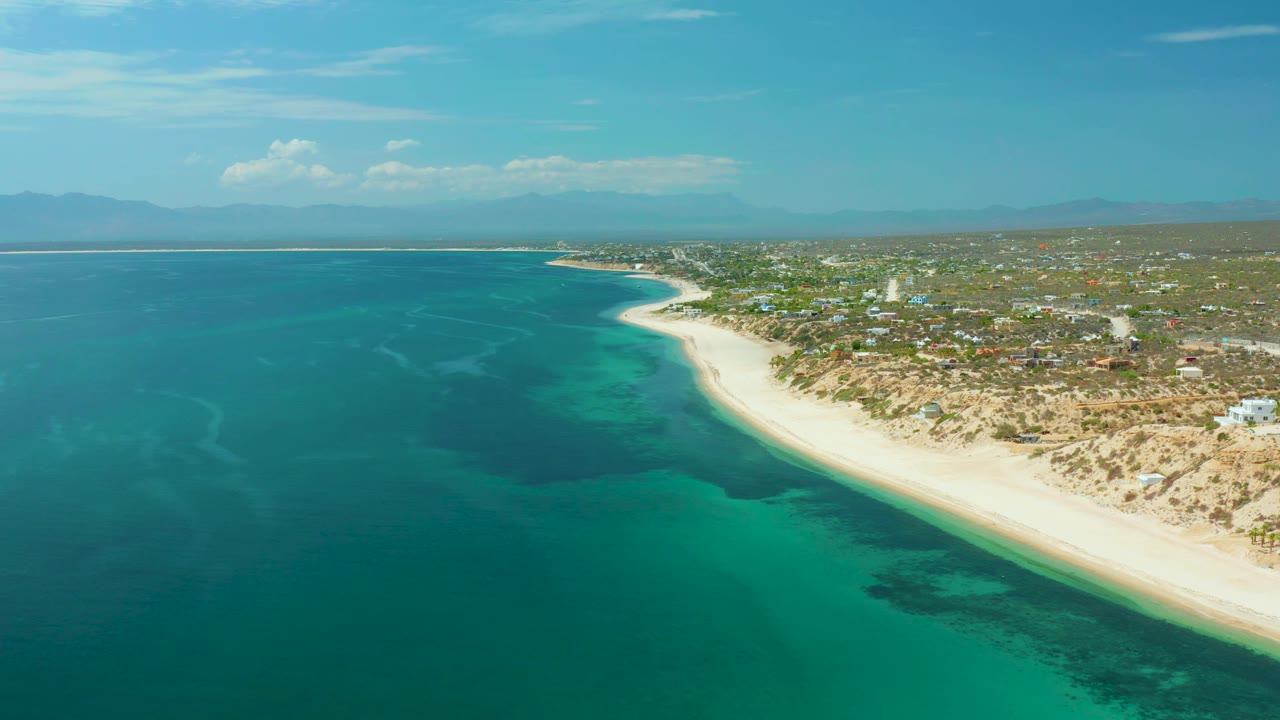 Aerial footage over El Sargento La Ventana, Mexico, shows turquoise shallows near the beach, darker water offshore, scattered buildings inland, and no visible people.