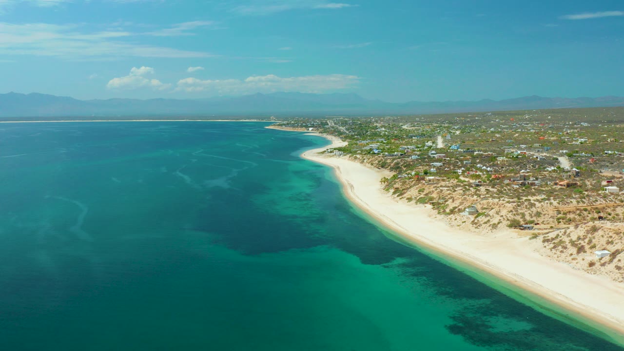 Elevated aerial frame over El Sargento La Ventana, Mexico, with turquoise water in the foreground, a curving sandy beach at right, inland vegetation and buildings beyond, and no visible people.