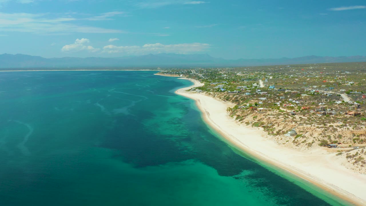 Opening aerial frame over El Sargento La Ventana, Mexico, with turquoise water, dark reef patches, a pale beach curve, low buildings inland, distant mountains, and no visible people.