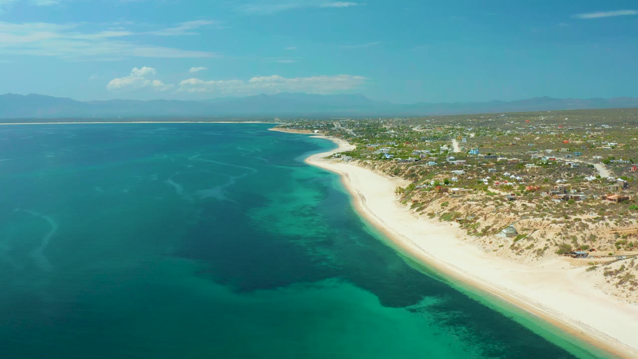 High drone view of blue-green water, pale shoreline, coastal houses, and mountains.