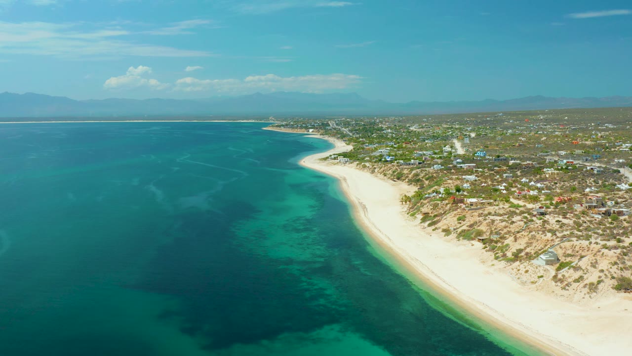 Drone frame over El Sargento La Ventana, Mexico, with emerald water on the left, surf edging the white sand, a spread of low buildings inland, hazy mountains beyond, and no visible people.